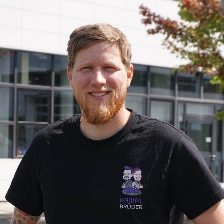 Lächelnder Mann mit blondem Bart und hellbraunem Shirt, vor hellem Hintergrund.